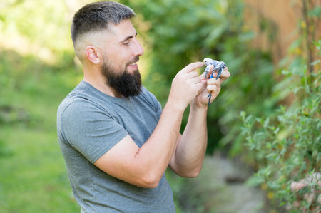 A bearded man with short hair holds a small bird in his hands. He's outdoors in a garden setting, sunlight illuminating the scene. The moment feels calm and tender.の写真素材