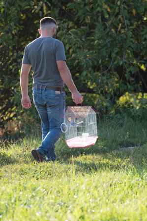 A man walks across a grassy area, carrying a small birdcage. The birdcage contains a small bird. The scene is peaceful and outdoors.の写真素材