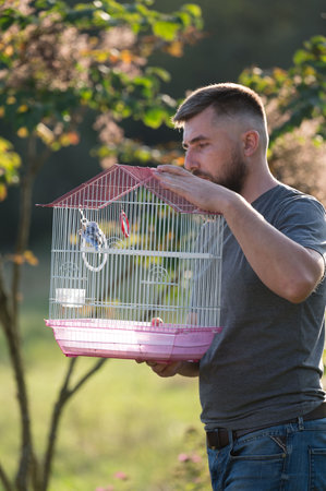 A man with a beard carefully carries a pink birdcage containing a small bird. He's standing outside in a sunny park setting, surrounded by green foliage.の写真素材