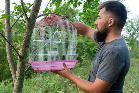 A bearded man carefully holds a pink and white birdcage containing a small bird. The setting is outdoors, amidst lush green foliage and a tree.の写真素材