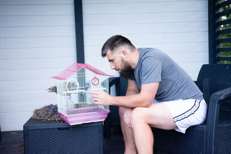 A bearded man sits on patio furniture, gently interacting with small birds inside a pink and white...