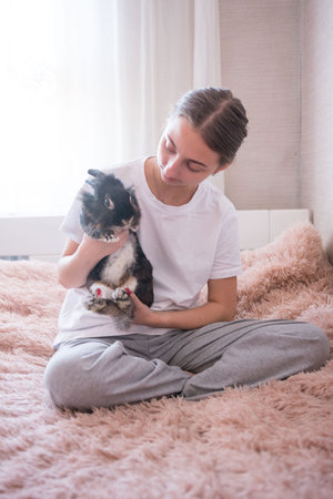 A young woman sits on a plush, pink bed, cradling a black and white rabbit. The scene is calm and intimate, highlighting the bond between human and pet.の写真素材