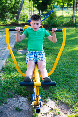 A young boy uses a yellow outdoor exercise machine, focused on his workout. He's wearing a green shirt and checkered shorts in a sunny park setting.の写真素材