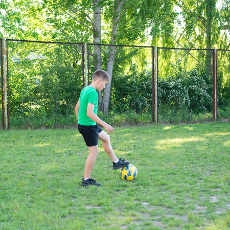 A boy in a green shirt and dark shorts practices his soccer skills outdoors. He juggles the ball with precision on a grassy area next to a chain-link fence. The scene is peaceful and sunny.の写真素材