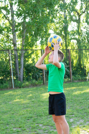 A boy in a green shirt and black shorts playfully balances a soccer ball on his head. He's outdoors on a grassy field, trees in the background.の写真素材
