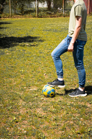 A young person in jeans and a t-shirt casually balances a soccer ball on their foot. The setting is outdoors on a grassy field on a bright day.の写真素材