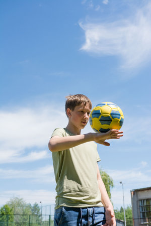 A teenage boy enjoys a sunny day by juggling a yellow and blue soccer ball. He's wearing a light green t-shirt and jeans. The background shows a clear blue sky and some greenery.の写真素材
