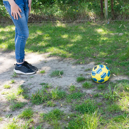 A child's feet near a yellow and blue soccer ball on a partly grassy, partly dirt ground. Sunlight dapples the scene.の写真素材