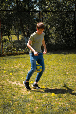 A young boy practices soccer in a sunny outdoor setting. He skillfully maneuvers a soccer ball through a series of cones. The background shows a grassy field and a wire fence.の写真素材