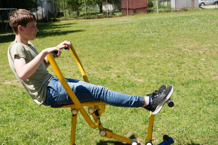 A young person enjoys a sunny day by exercising on a simple outdoor fitness machine. The machine is yellow and the grass is green, showing a vibrant setting.の写真素材