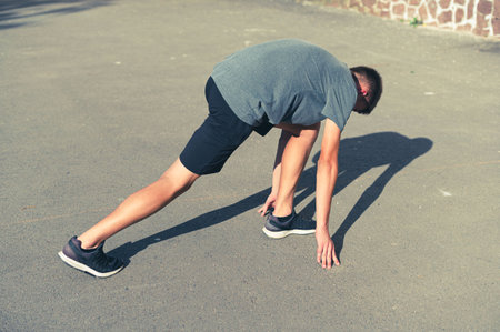 A young man in athletic wear performs a runner's stretch on asphalt. Sunlight casts shadows on the ground. He's focused and ready for physical activity.の写真素材