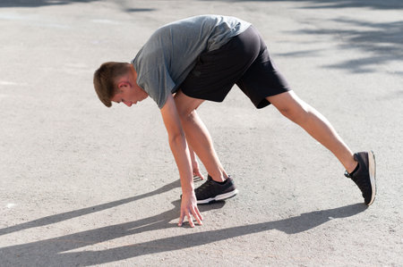 A teenage boy performs a dynamic stretch on an asphalt surface. He's wearing athletic clothing and...