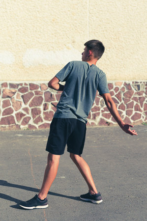 A young man, seen from behind, is playing a game outdoors. He's wearing athletic clothing and is focused on his throw near a stone wall.の写真素材