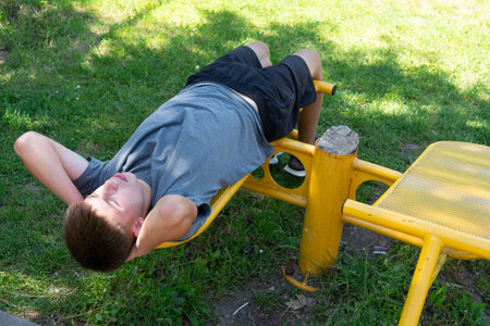 A teenage boy lies on a yellow outdoor fitness bench, taking a break after exercising. He's wearing a gray shirt and dark shorts. The scene is set in a park on a sunny day.の写真素材