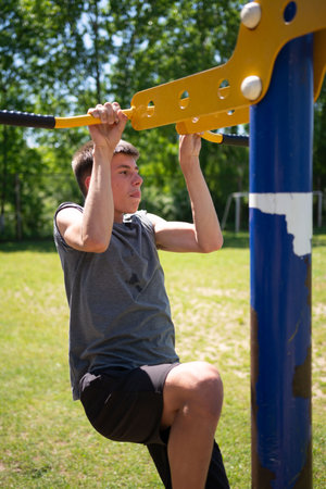 A teenage boy performs pull-ups on outdoor exercise equipment. He's wearing a gray shirt and dark...