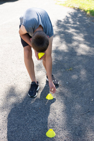 A young person bends down, placing yellow training cones on the ground. They're likely preparing for an exercise session outdoors on a sunny day.の写真素材