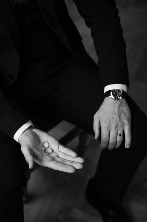 Close-up of a man in a suit gently holding two wedding bands. The image is black and white,...