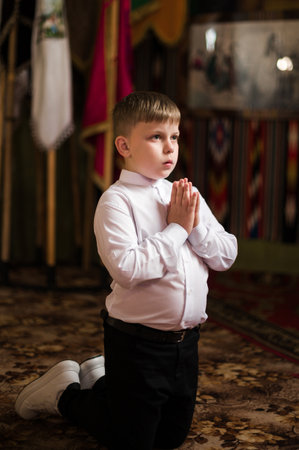 A young boy, dressed in a white shirt and dark pants, kneels in prayer with his hands clasped. The background suggests a place of worship or quiet contemplation.の写真素材