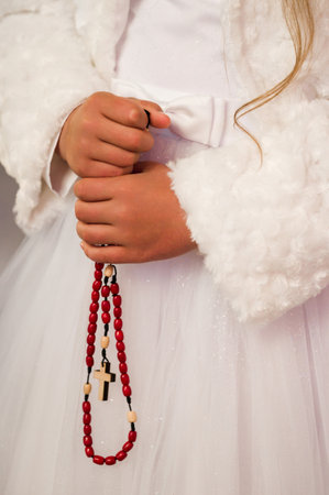 Close-up of a young girl in a white dress, gently holding a red beaded rosary. The scene evokes a sense of peace and religious devotion.の写真素材