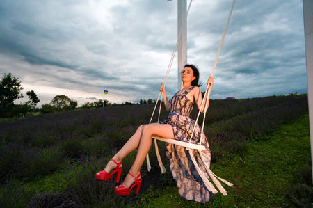 A serene moment captured: a woman in a flowing dress sits on a swing, surrounded by a lavender field. The sky is overcast, adding to the peaceful atmosphere.の写真素材