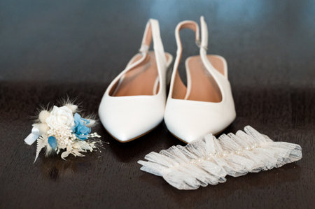 Close-up of white bridal shoes, a delicate lace garter, and a small, pretty flower boutonniere arranged on a dark surface. Wedding details.の写真素材