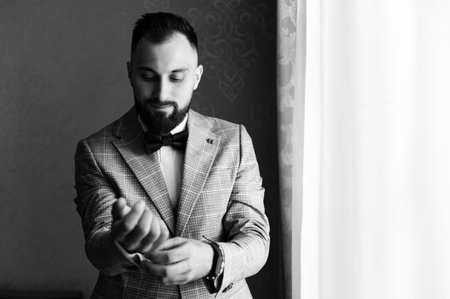 A black and white portrait of a groom in a suit, meticulously adjusting his cufflinks. He stands...