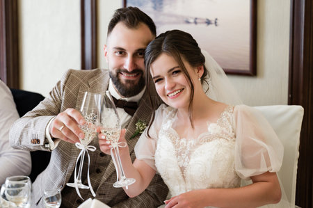 A newly married couple joyfully raises their champagne flutes in a toast. The bride wears a beautiful white dress, and the groom is in a brown suit. Pure happiness radiates from the image.の写真素材