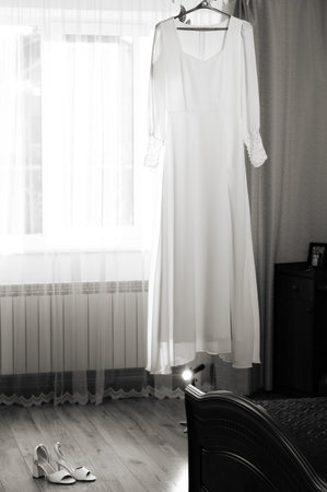 A white wedding dress hangs near a window, bathed in soft light. A pair of elegant bridal shoes rests on the floor nearby, adding to the serene atmosphere of the preparation.の写真素材