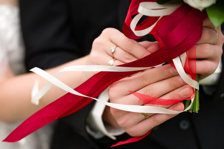 Close-up of a bride and groom's hands gently holding a wedding bouquet. Red and white ribbons elegantly intertwine, adding to the romantic atmosphere.の写真素材