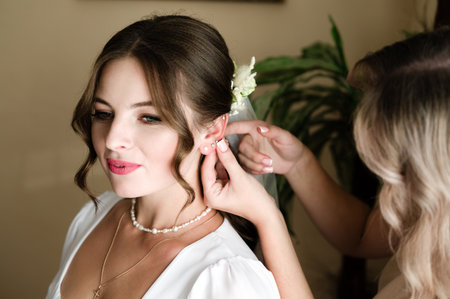 A bride is getting ready for her wedding. A friend gently places earrings in her ears. The bride...