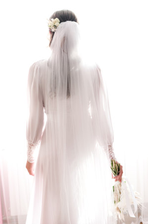 A bride stands before a window, her back to the camera. She wears a long white dress and veil, holding a delicate bouquet. Sunlight illuminates her figure.の写真素材
