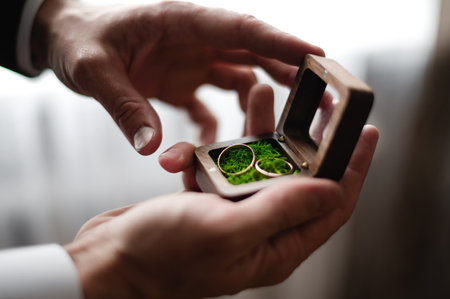 A person delicately holds a small, wooden box containing two gold wedding bands. The rings rest on a bed of green moss, adding a touch of nature to the scene.の写真素材