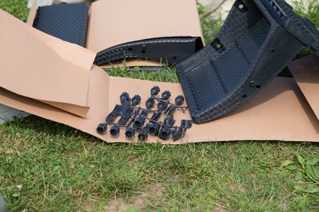 Black plastic furniture parts and small black fasteners lie on brown cardboard on a grassy surface. Looks like unassembled outdoor furniture.の写真素材