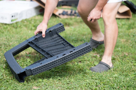 Close-up of a person assembling a piece of outdoor furniture. The dark gray plastic component is being carefully positioned on a grassy lawn.の写真素材
