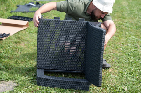 A person assembles dark gray wicker-style outdoor furniture on a grassy lawn. The pieces appear to be part of a seating set.の写真素材