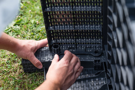 Close-up of hands assembling dark gray plastic components. The structure appears modular, with interlocking parts. Set against a backdrop of green grass.の写真素材