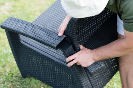 A person assembles a dark-colored wicker chair outdoors. The chair is made of woven plastic and appears sturdy. The setting is a grassy lawn.の写真素材