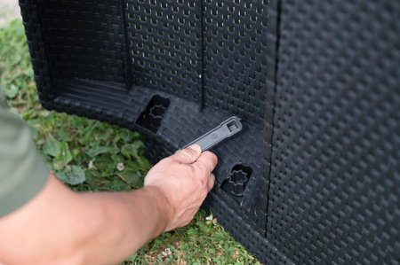 Close-up of hands assembling dark-colored woven plastic outdoor furniture. A small plastic tool is used to connect the pieces together. The setting is outdoors on grass.の写真素材