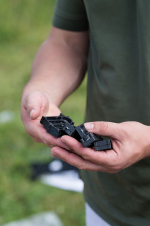 Close-up of hands holding several small, black plastic pieces. The person is wearing a dark green shirt and is outdoors in a grassy area.の写真素材
