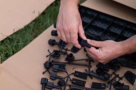 A person assembles black plastic components outdoors. The parts are scattered on cardboard, set against a grassy backdrop. Focus is on the hands connecting two pieces.の写真素材