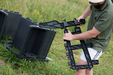 A person assembles a dark-grey plastic frame outdoors on grass. The parts interlock easily. Nearby are other similar pieces, likely for a patio chair or set.の写真素材