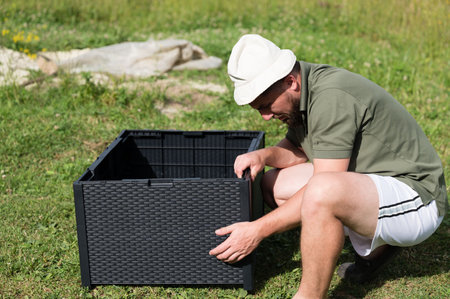 A man assembles a dark gray plastic raised garden bed in a grassy area. He is squatting down, carefully connecting the pieces. The setting is sunny and outdoors.の写真素材