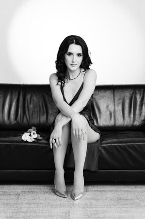 A stylish woman in a dark dress sits gracefully on a black leather sofa. A small bouquet rests beside her. The image is in black and white.の写真素材