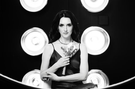 A black and white portrait of a woman with long dark hair, wearing a satin slip dress and holding a small bouquet. The background features circular lights.の写真素材