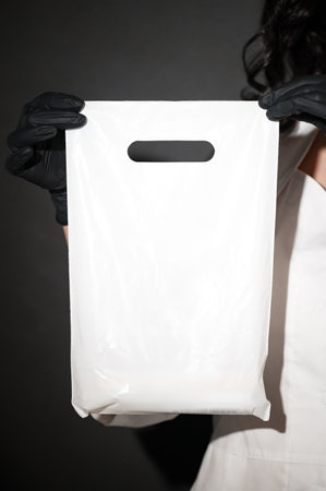 Close-up of a hand in a black glove holding a plain white shopping bag. The bag features contact information, ideal for business promotion.の写真素材