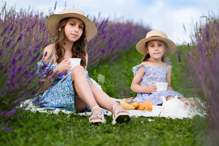Two girls in summer dresses and straw hats have a picnic in a beautiful lavender field. They sip from teacups, enjoying the idyllic setting. Fruit and a teapot complete the scene.の写真素材