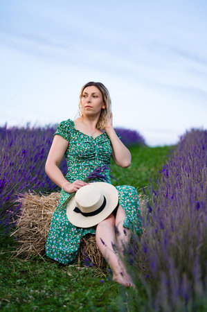A serene scene of a woman in a green floral dress, sitting on a hay bale amidst a vibrant lavender field. She holds a straw hat, creating a peaceful and idyllic atmosphere.の写真素材