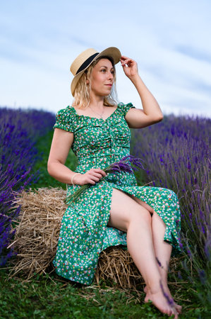 A woman in a green floral dress and straw hat sits on a hay bale amidst a vibrant lavender field. She holds a small bouquet of lavender, enjoying the summer scenery.の写真素材