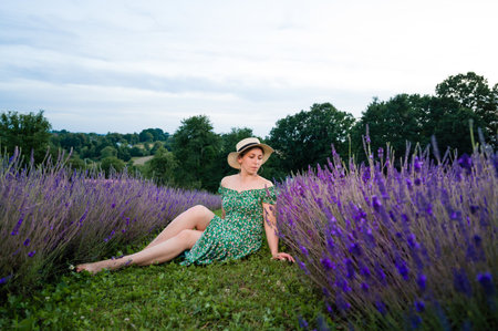 A serene scene of a woman sitting amidst rows of vibrant purple lavender flowers. She wears a floral dress and straw hat, enjoying the tranquil countryside view.の写真素材