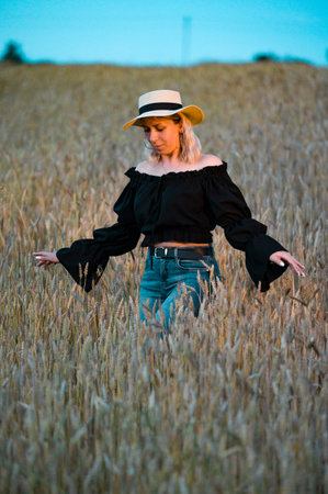 A blonde woman strolls through a golden wheat field during the evening. She's dressed in a black off-the-shoulder top and blue jeans. The sunset casts a warm glow.の写真素材